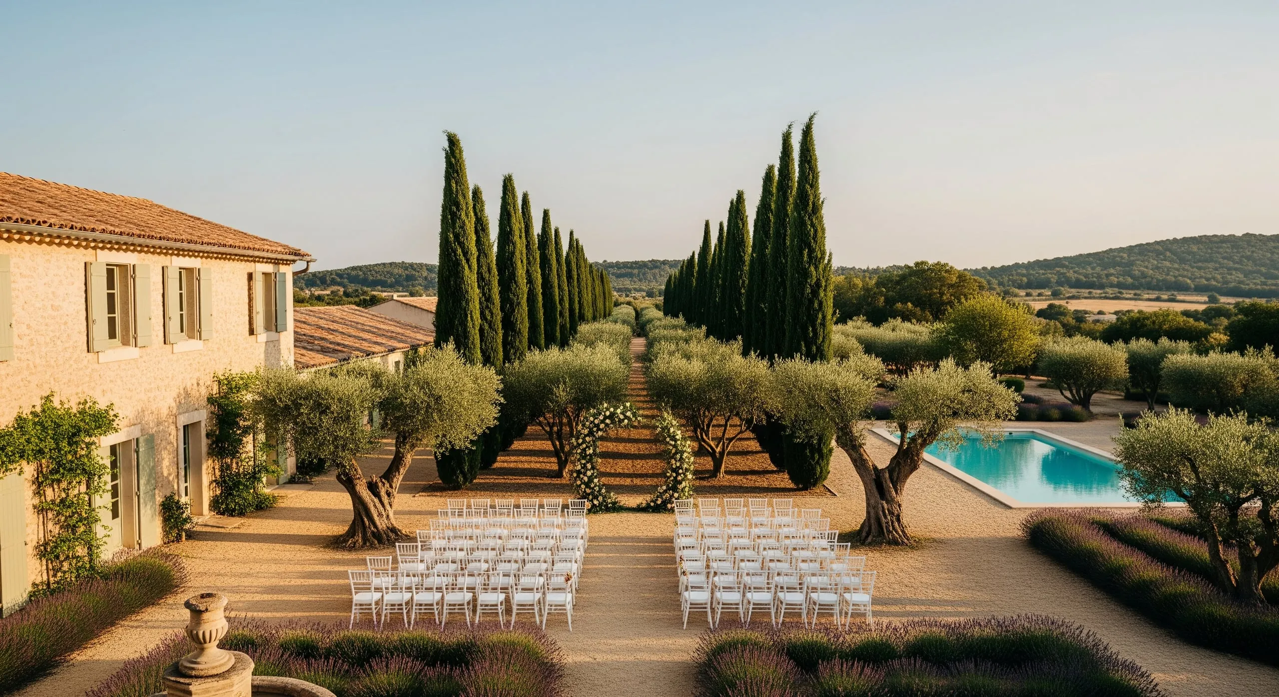 Cérémonie laïque dans l'oliveraie : arche fleurie de roses blanches entre les cyprès, rangées de chaises blanches face aux collines du Luberon.